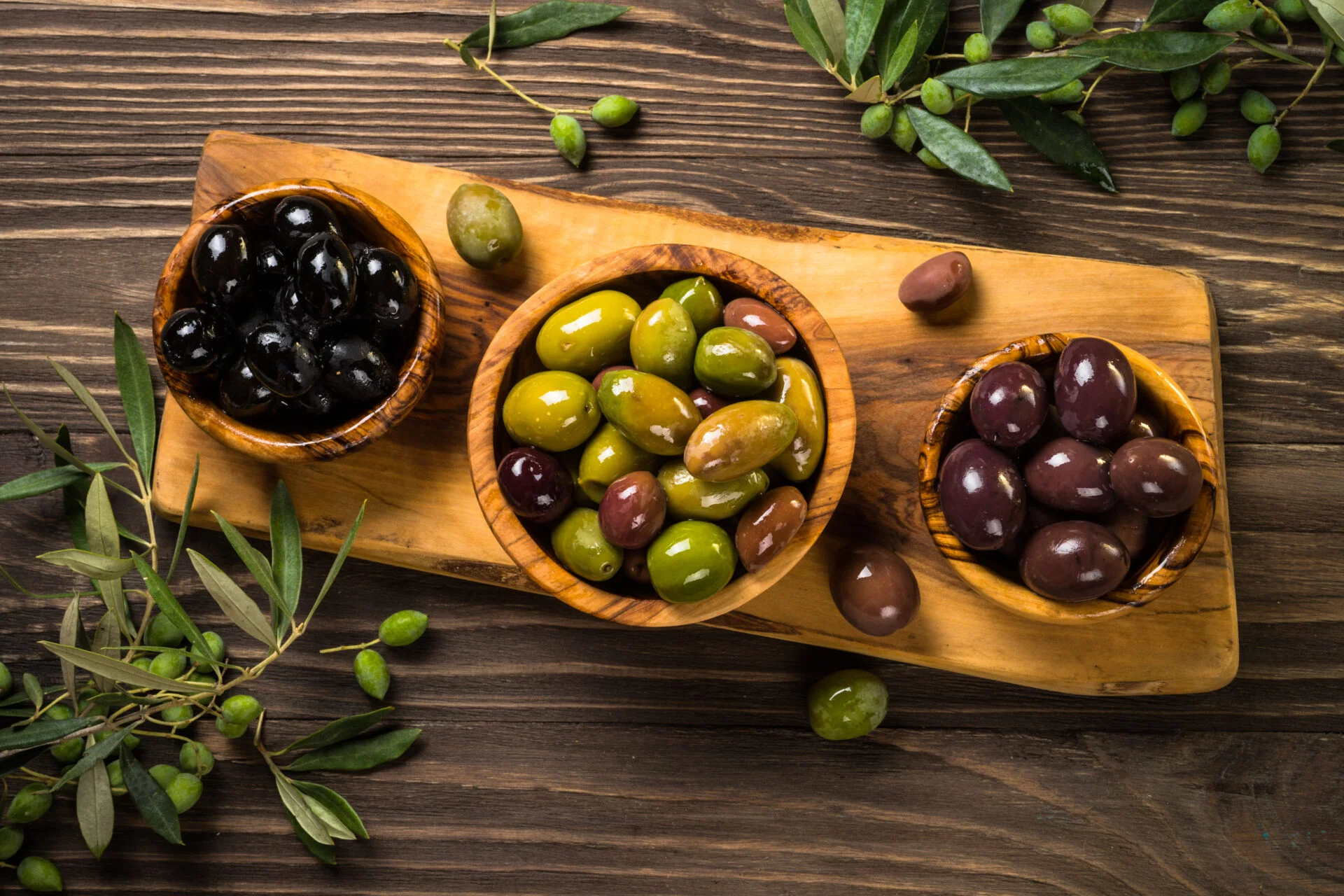 Black and green olives in wooden bowls with natural olive leaves on dark wooden table. Top view.