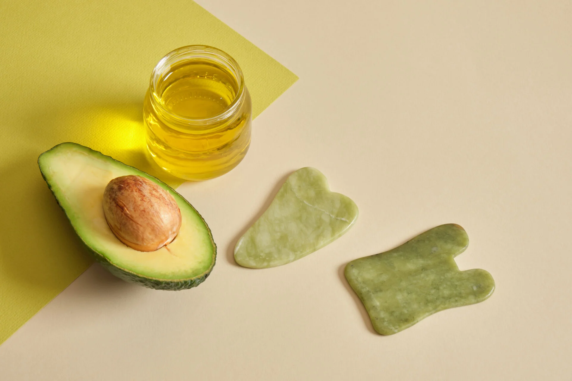 massage with avocado oil concept, avocado oil in a jar, sliced avocados and gua sha slices on a green and beige background