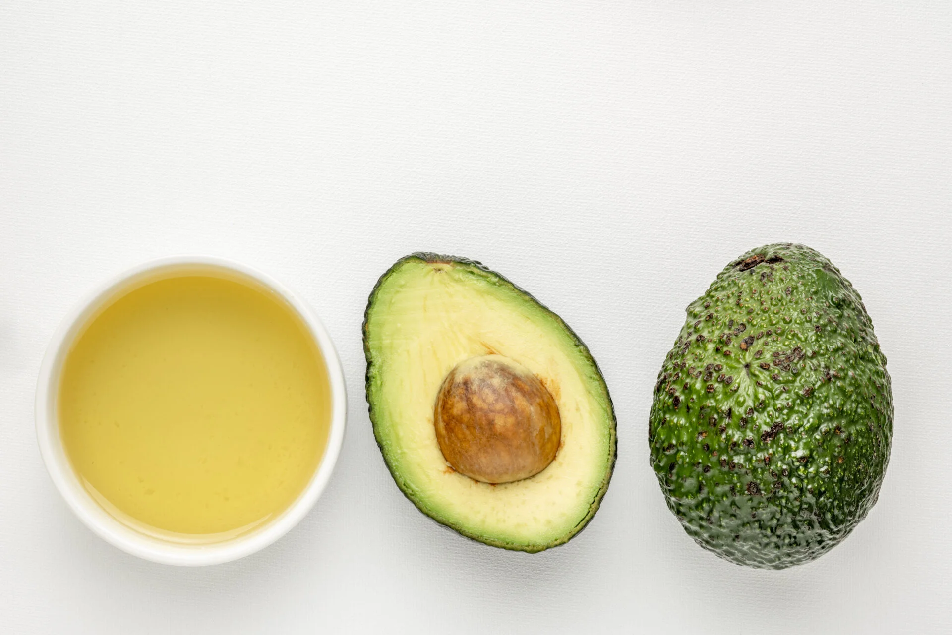 avocado oil in a small ceramic bowl and avacado fruit against white art canvas