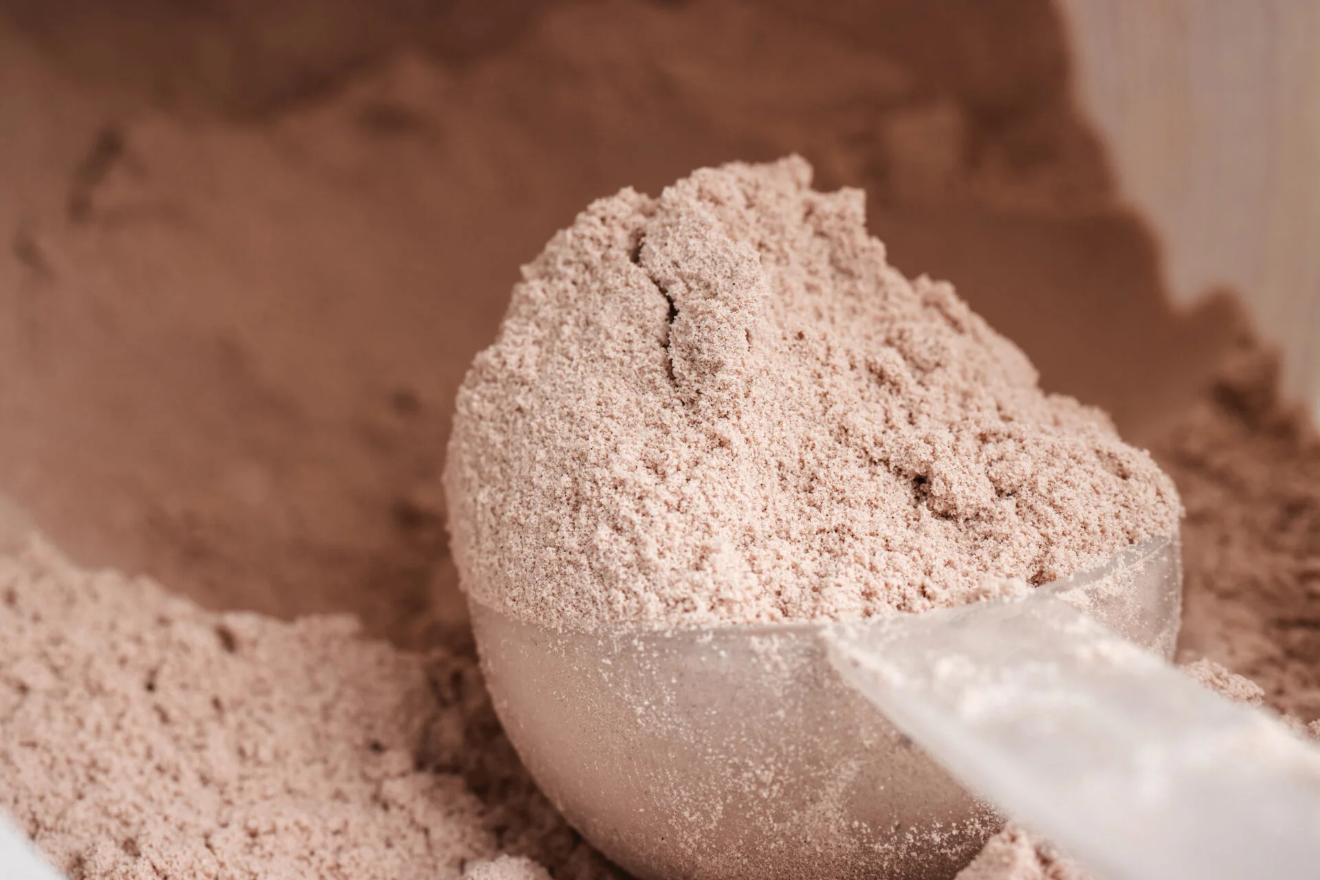 Scoop of chocolate whey protein isolate in a plastic jar, close-up view.