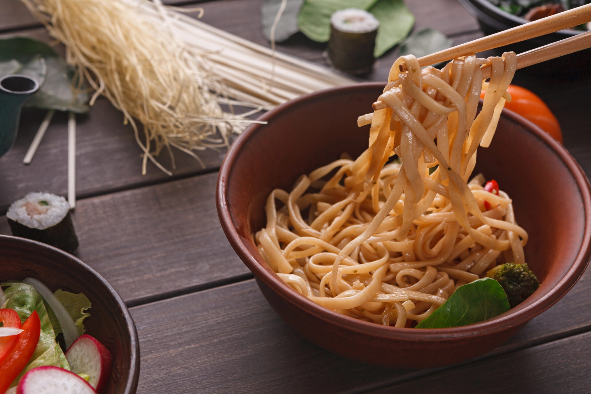 Chopsticks, taking rice noodles with soy sauce in bowl on wooden background, close-up